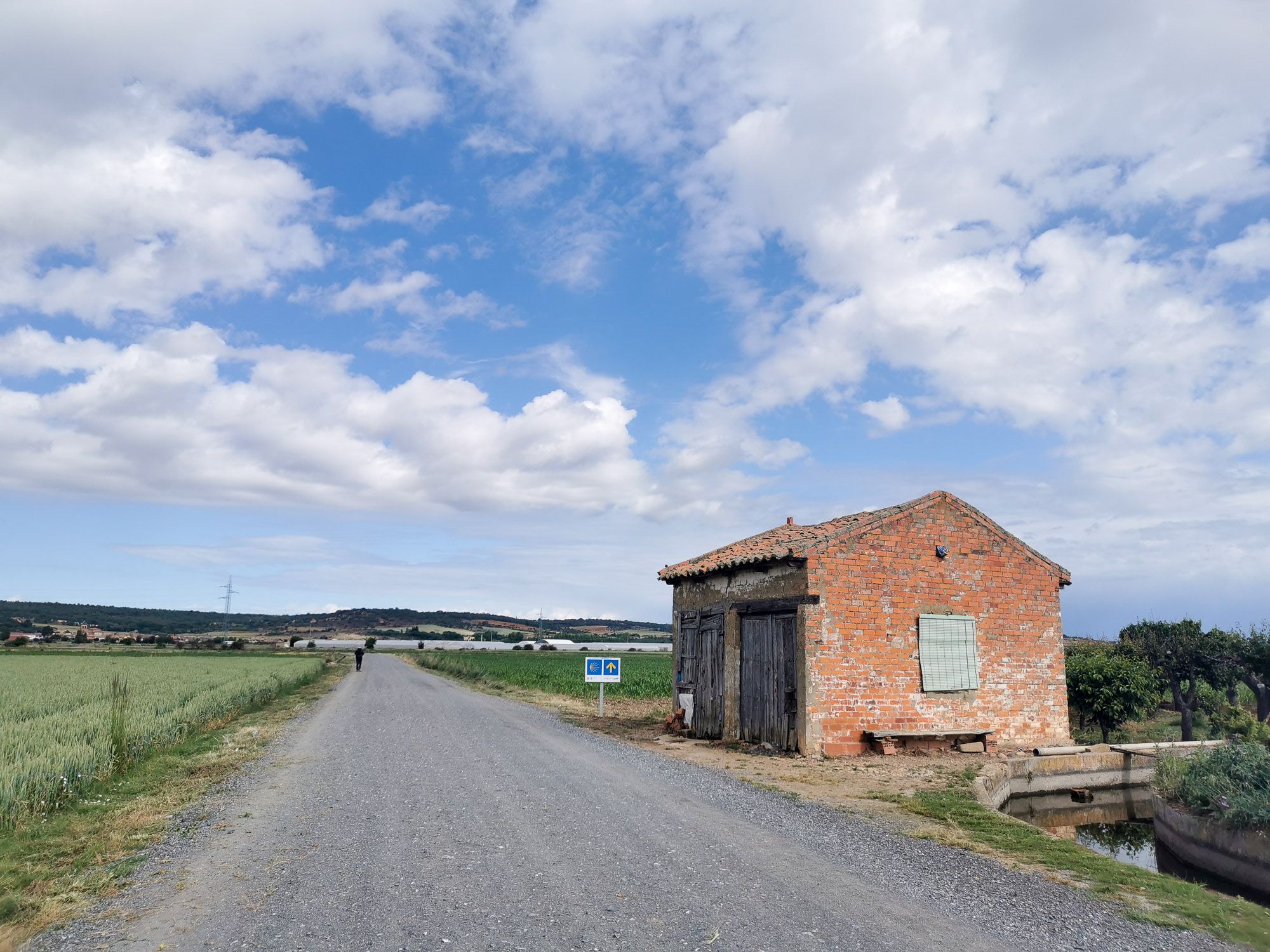 ทางเดินออกจากเมือง Hospital de Órbigo ทางเดินเป็นถนนกรวด ด้านข้างเป็นทุ่งเกษตรกรรม มีสิ่งก่อสร้างอิฐ และป้ายชี้ทาง Camino อยู่ทางขวา