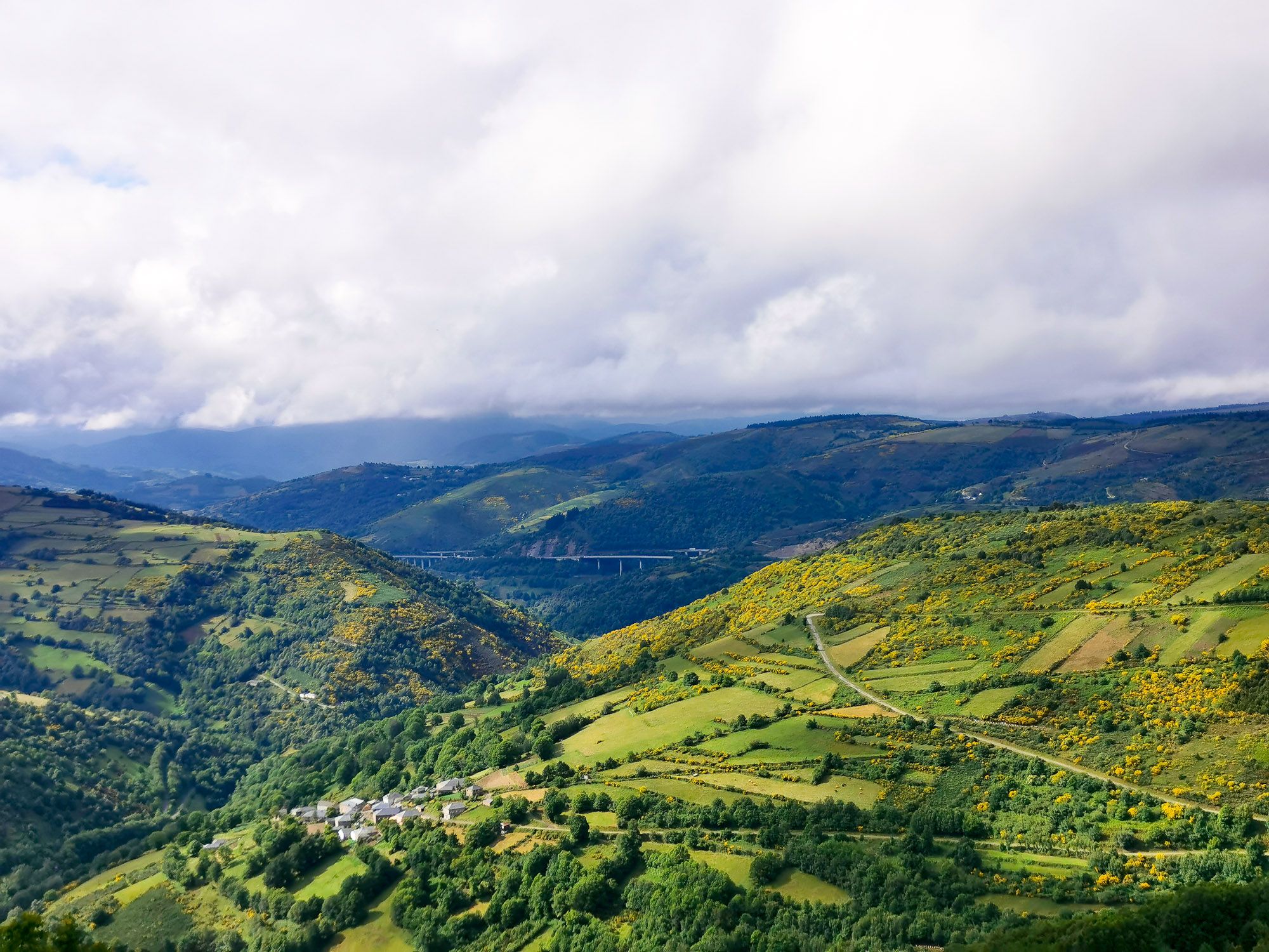 เทือกเขาลดหลั่นกัน มีหมู่บ้านเห็นไกล ๆ และดอกไม้สีเหลือง สลับกับทุ่งหญ้าสีเขียวที่เนินเขา