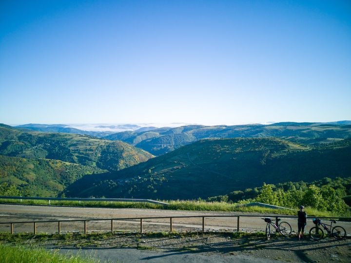 วิวจากหมู่บ้าน O Cebreiro เป็นเทือกเขาซ้อนทับกันไปมา มีเมฆลอยต่ำด้านหลัง ด้านหน้ามีคนจอดจักรยานชมวิว